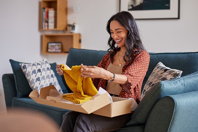 Femme souriante qui déballe un vêtement jaune d’un carton sur son canapé